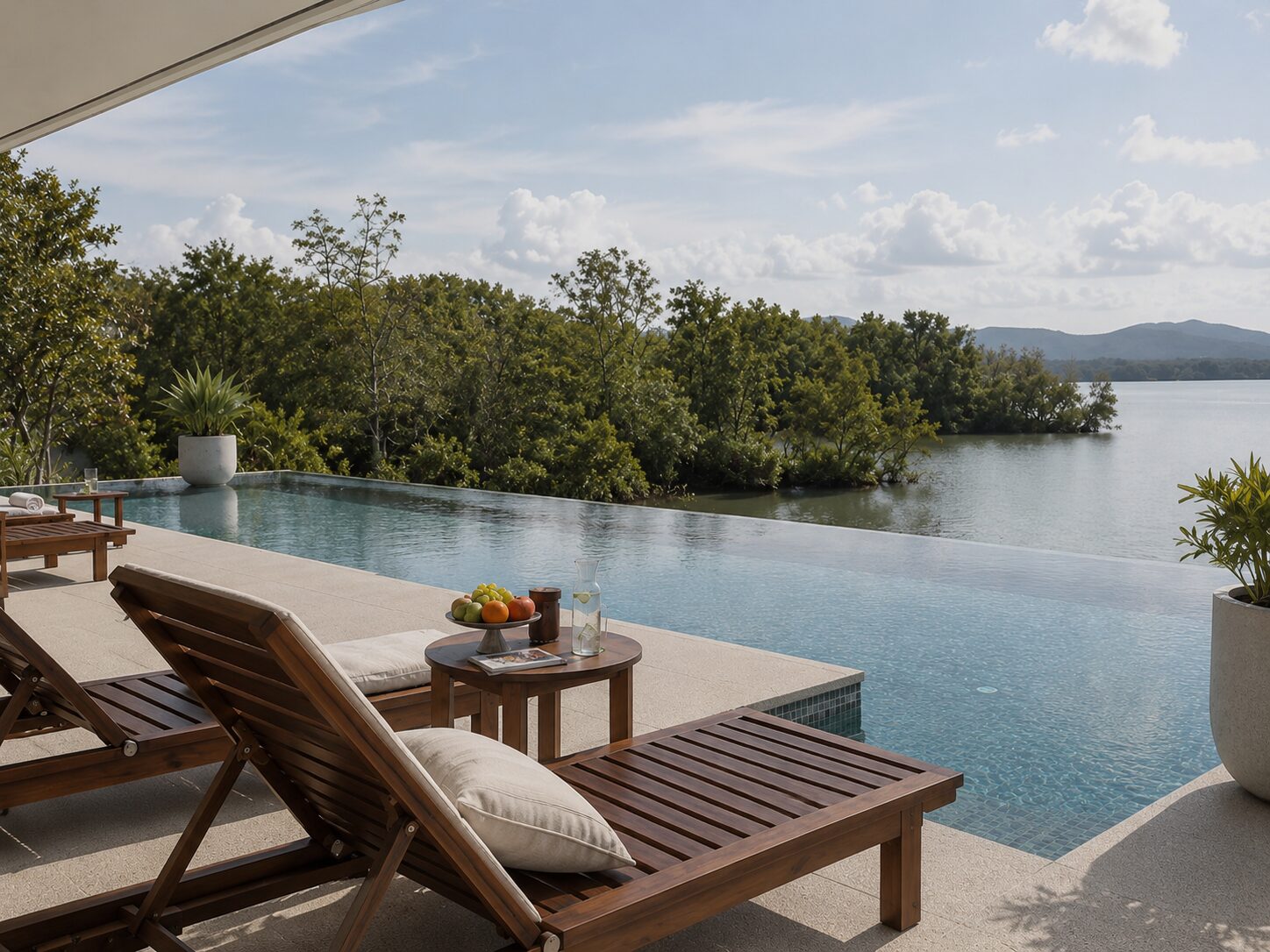Infinity pool overlooking a lake with trees and distant hills; wooden lounge chairs and a small table with fruit appetizingly arranged.