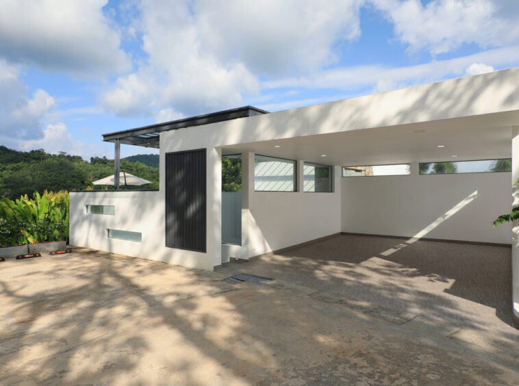 Modern white building with a covered carport and large rectangular windows overlooking a driveway, hillside and blue sky with clouds in the background
