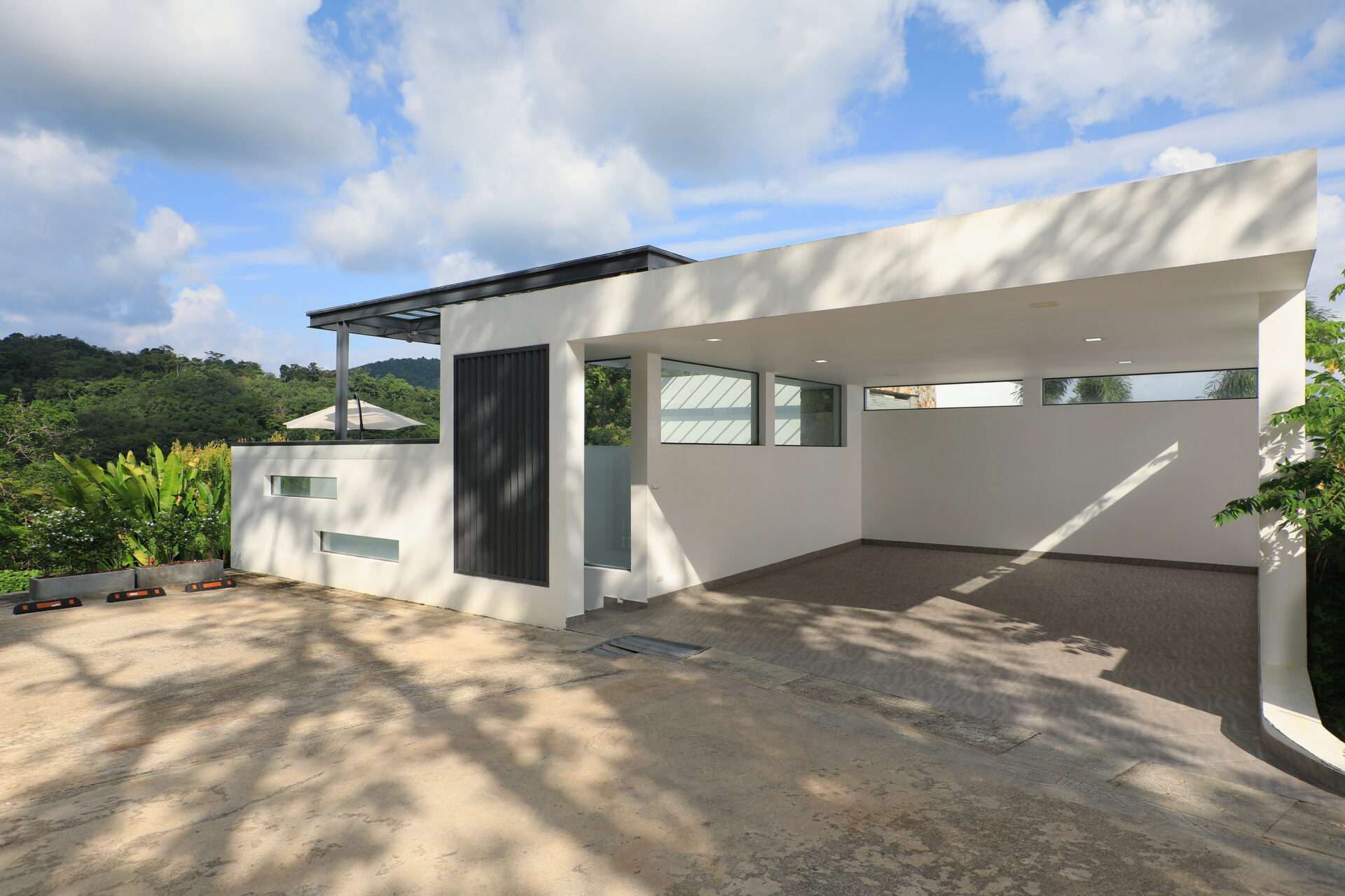 Modern white building with a covered carport and large rectangular windows overlooking a driveway, hillside and blue sky with clouds in the background