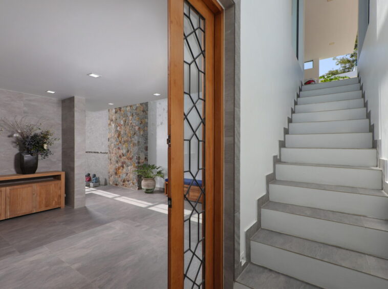 Modern foyer with a wooden-framed glass door, gray tile floor, and a white staircase to the right beside a stone accent wall and plants.