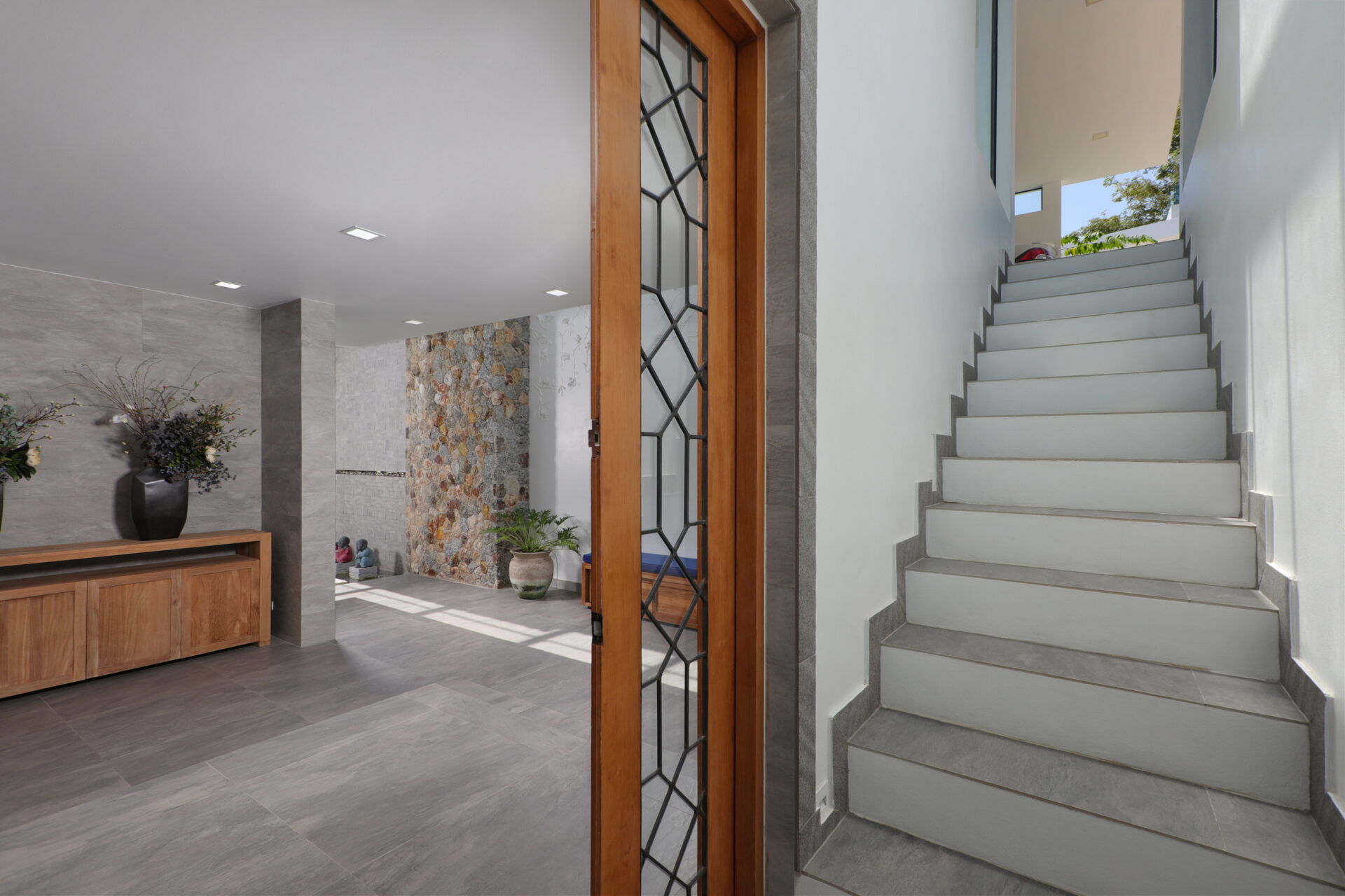 Modern foyer with a wooden-framed glass door, gray tile floor, and a white staircase to the right beside a stone accent wall and plants.
