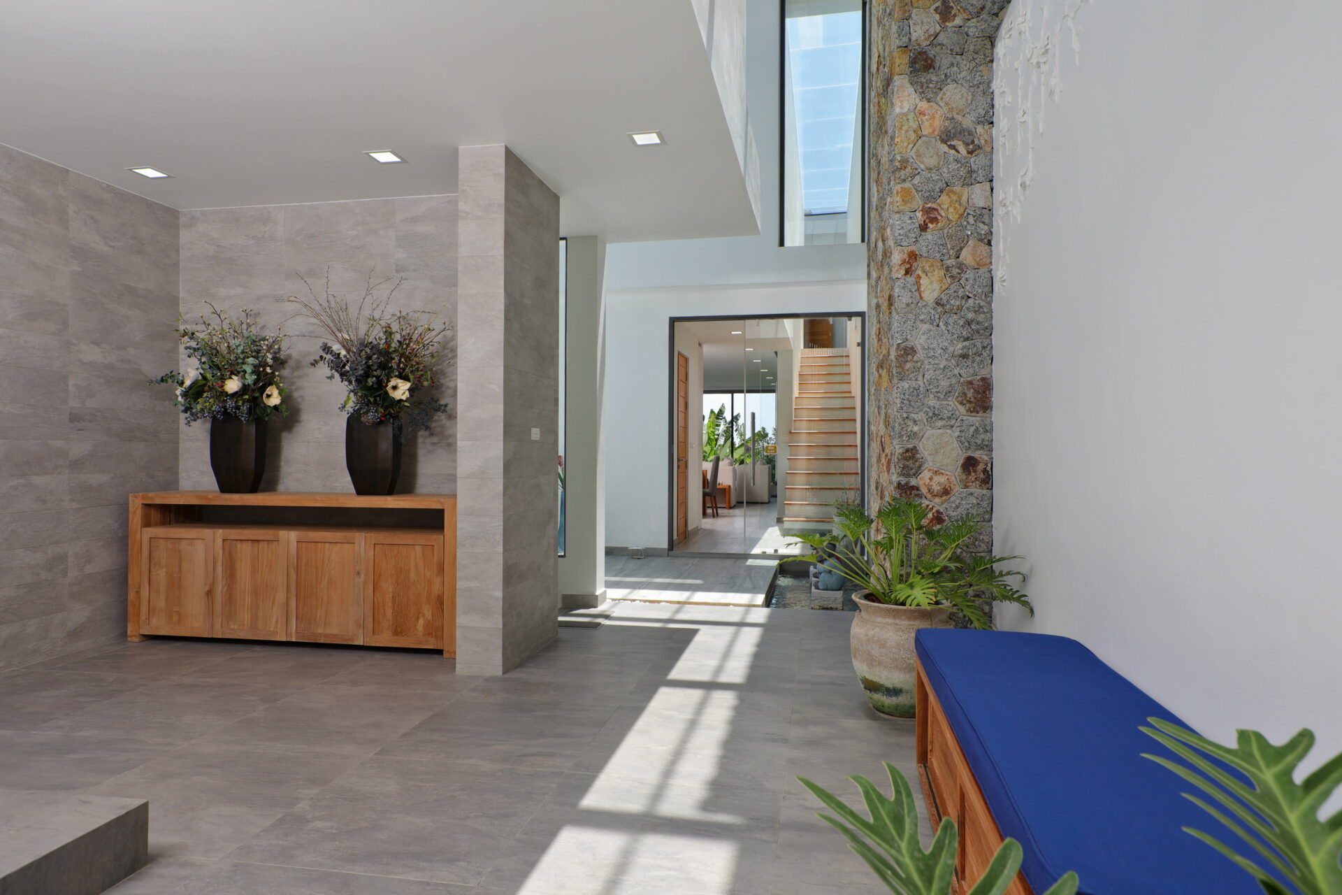 Modern lobby with gray tile walls, a wooden cabinet flanked by two black vases of flowers, and a stone wall on the right.