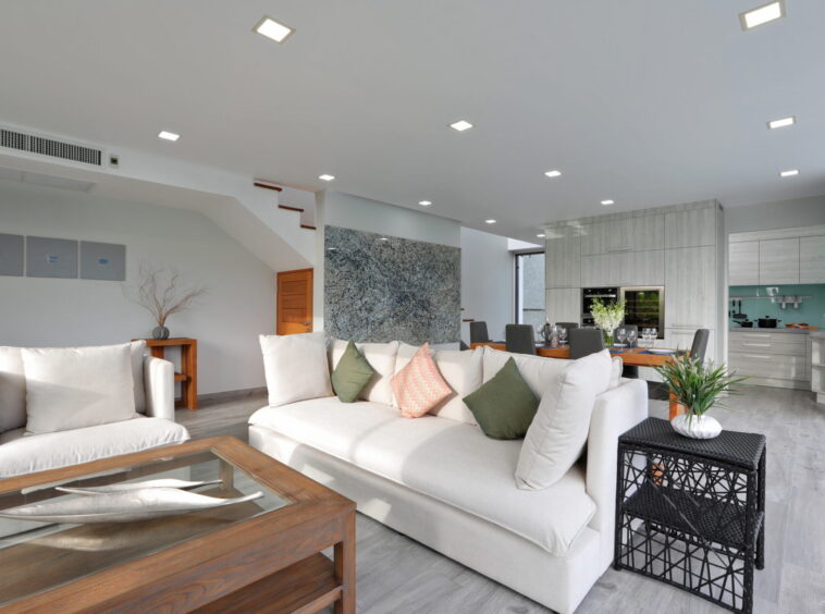 Open-plan living area with a light gray sectional sofa, wooden coffee table, and a modern kitchen in the background.