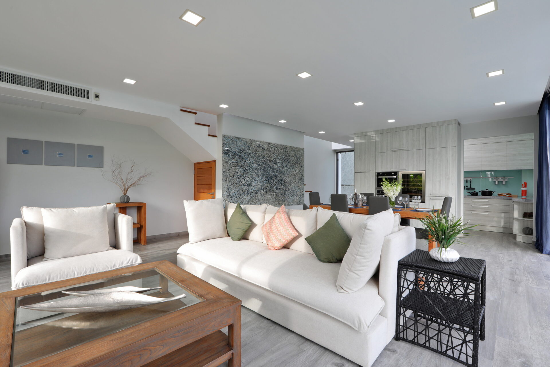 Open-plan living area with a light gray sectional sofa, wooden coffee table, and a modern kitchen in the background.