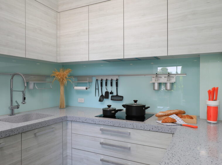 Modern kitchen with light wood cabinets, teal backsplash, stainless faucet, and utensils on a rail above the counter; pots on the stove and bread on a cutting board.