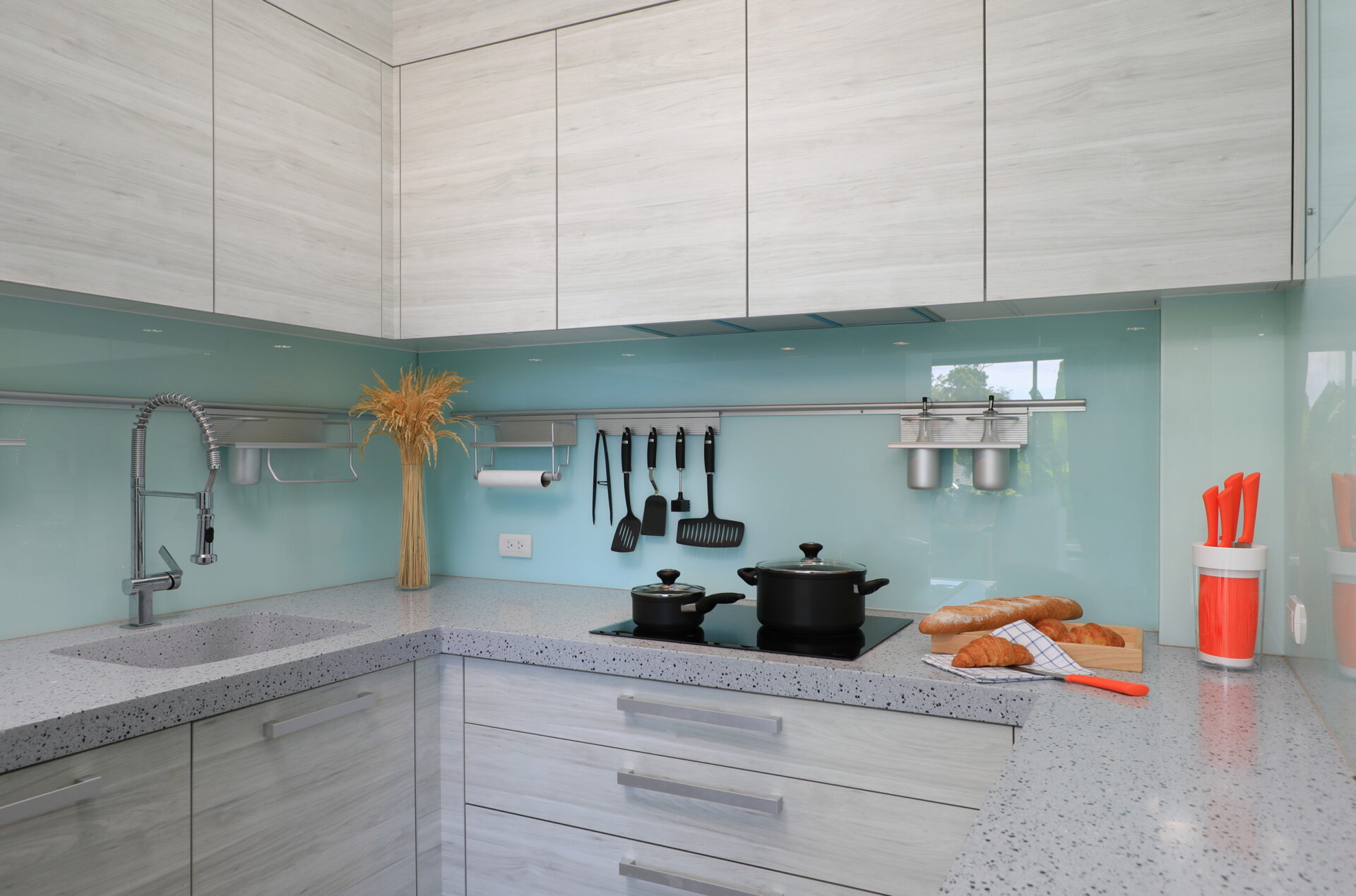 Modern kitchen with light wood cabinets, teal backsplash, stainless faucet, and utensils on a rail above the counter; pots on the stove and bread on a cutting board.