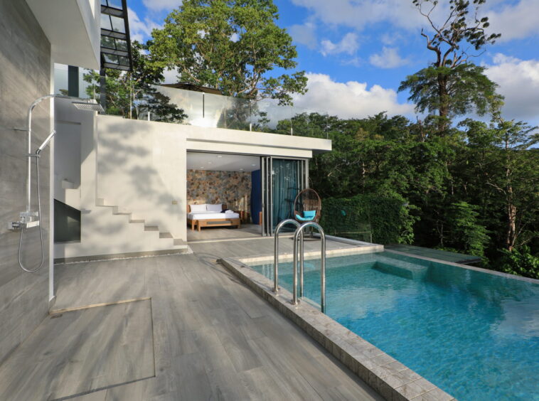 Modern poolside patio of a contemporary house with glass railing, outdoor shower, and lounging area overlooking a green forest.