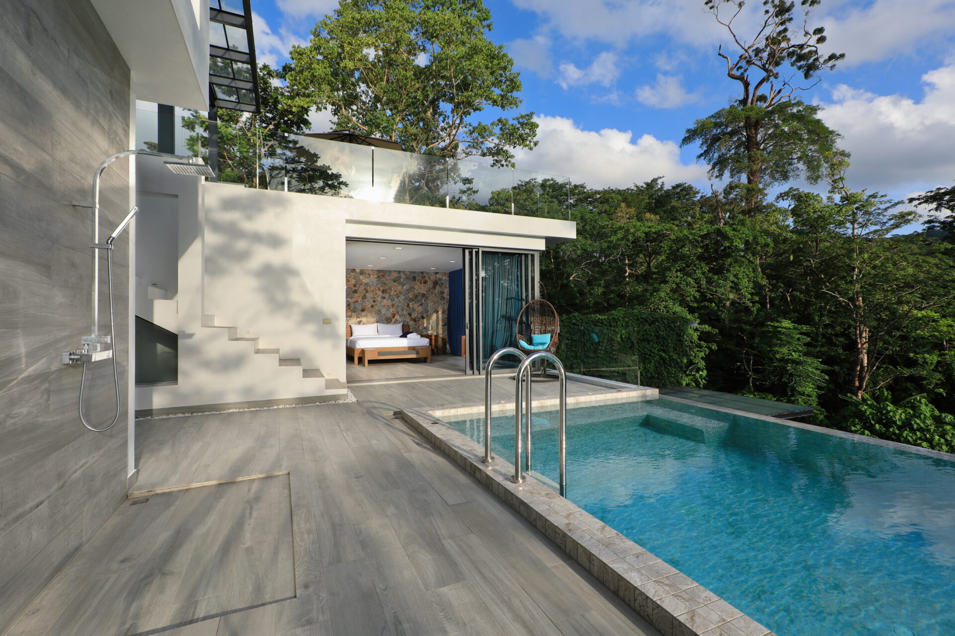 Modern poolside patio of a contemporary house with glass railing, outdoor shower, and lounging area overlooking a green forest.