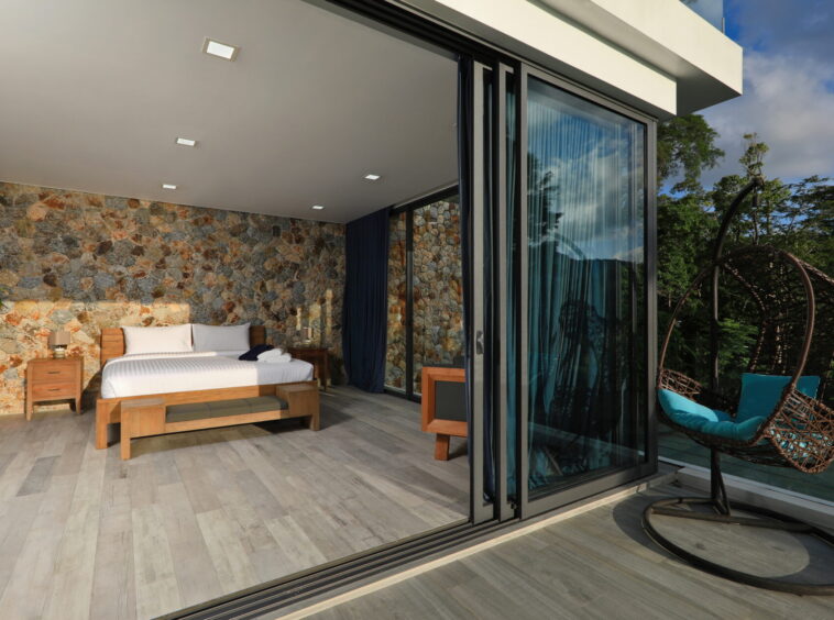 Hotel bedroom with stone accent wall, wooden bed, and side tables opening to a glass balcony door.
