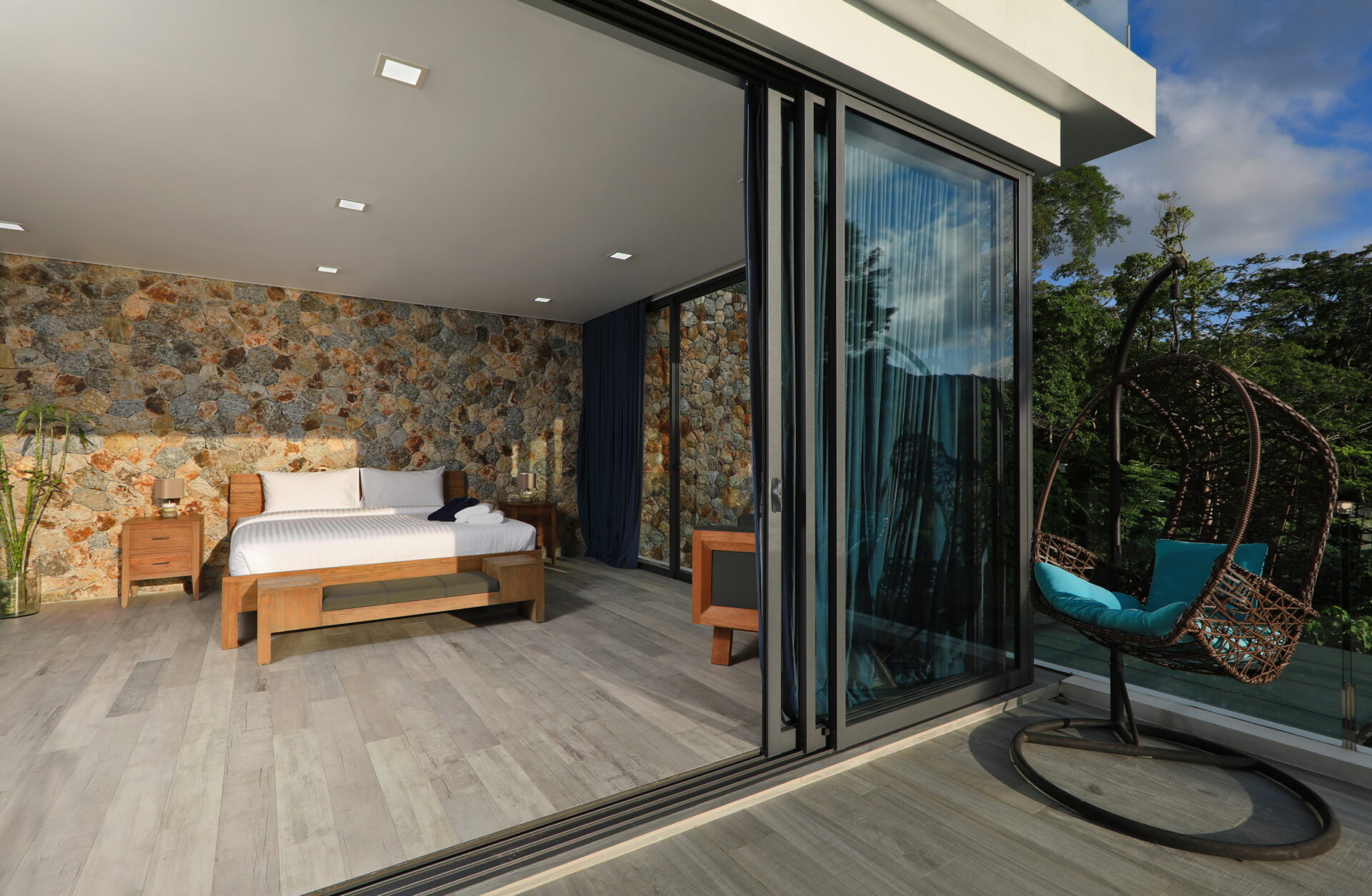 Hotel bedroom with stone accent wall, wooden bed, and side tables opening to a glass balcony door.