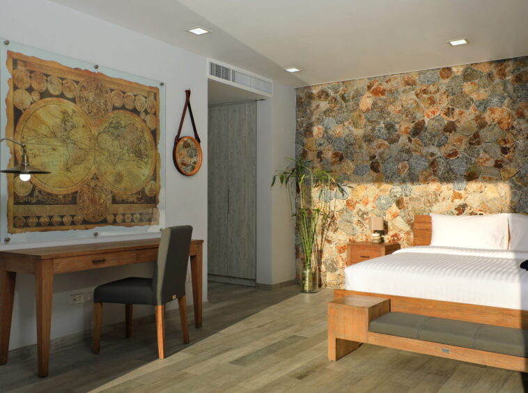 Hotel room with a large world map artwork, wooden desk and chair, and a white‑lined bed against a stone accent wall