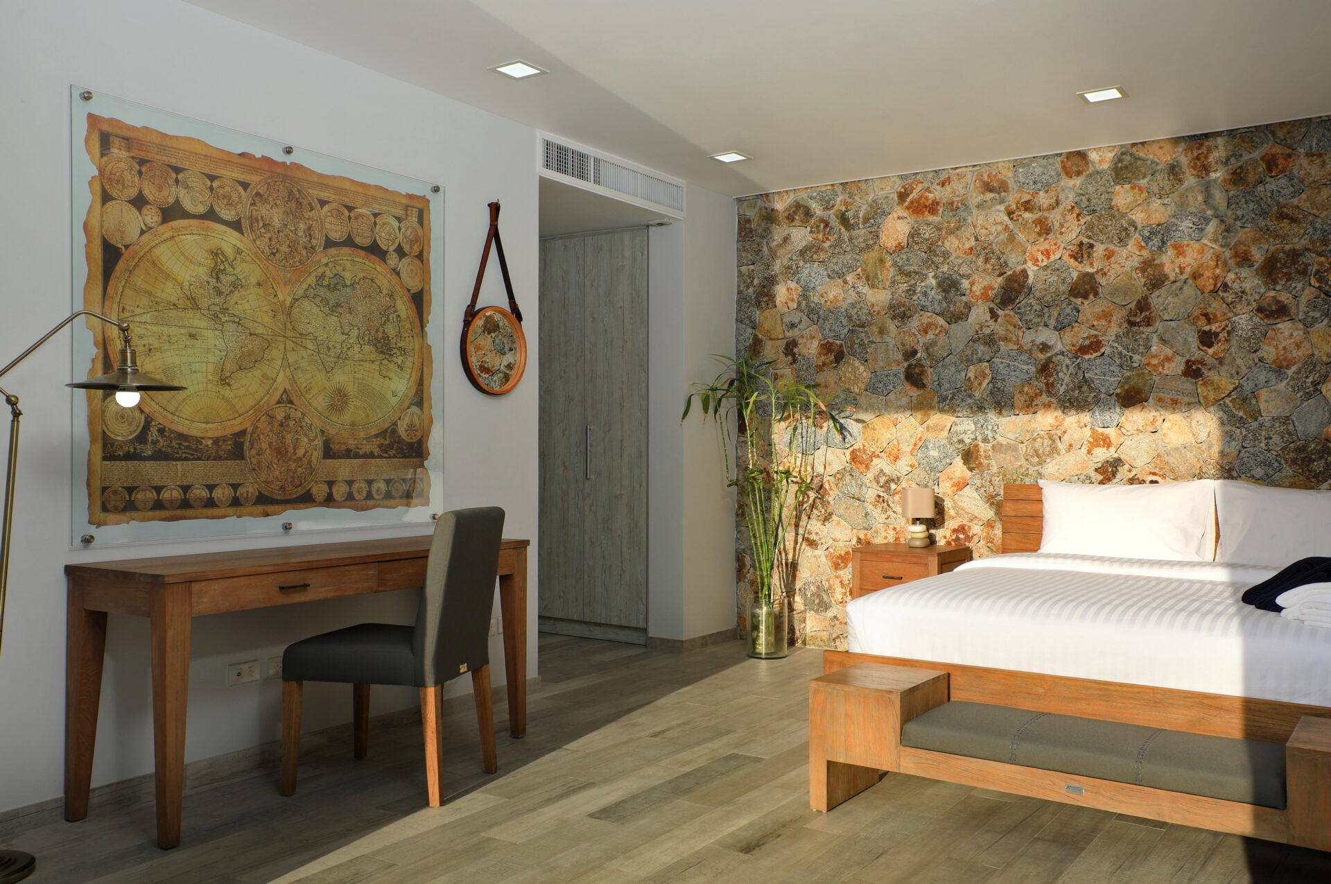 Hotel room with a large world map artwork, wooden desk and chair, and a white‑lined bed against a stone accent wall
