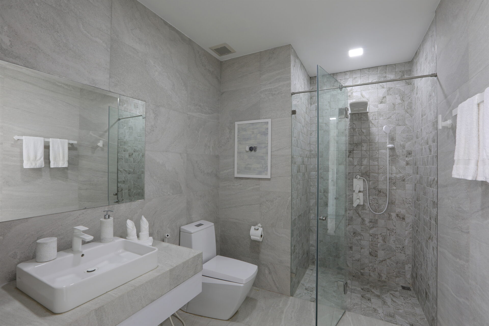 Gray marble bathroom with a wide rectangular sink, wall mirror, toilet, and a glass-enclosed shower area.