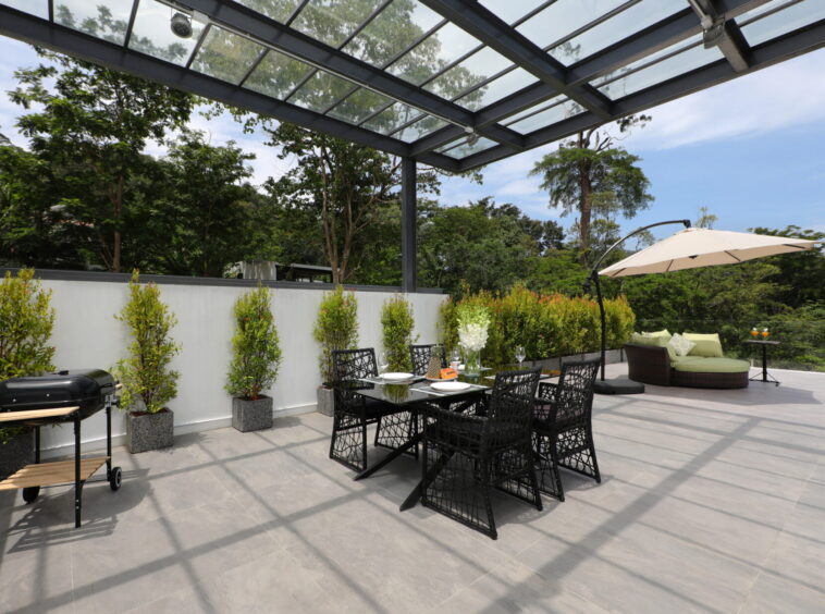 Modern outdoor patio with a glass-covered pergola, a black dining table and chairs, potted shrubs along a white wall, and a beige umbrella edge-to-right.