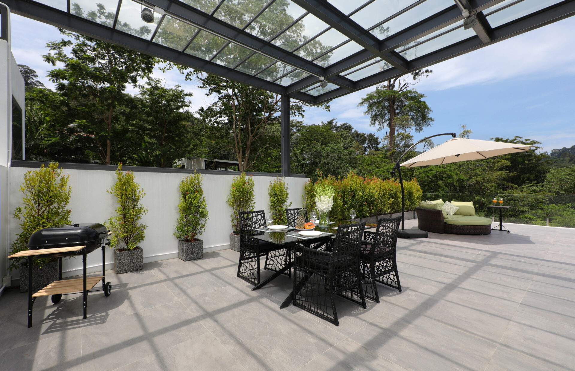 Modern outdoor patio with a glass-covered pergola, a black dining table and chairs, potted shrubs along a white wall, and a beige umbrella edge-to-right.