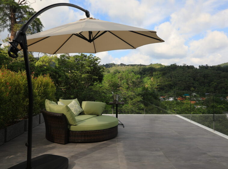 Outdoor terrace featuring a circular wicker sofa with green cushions under a beige cantilever umbrella, overlooking hills and a blue sky.