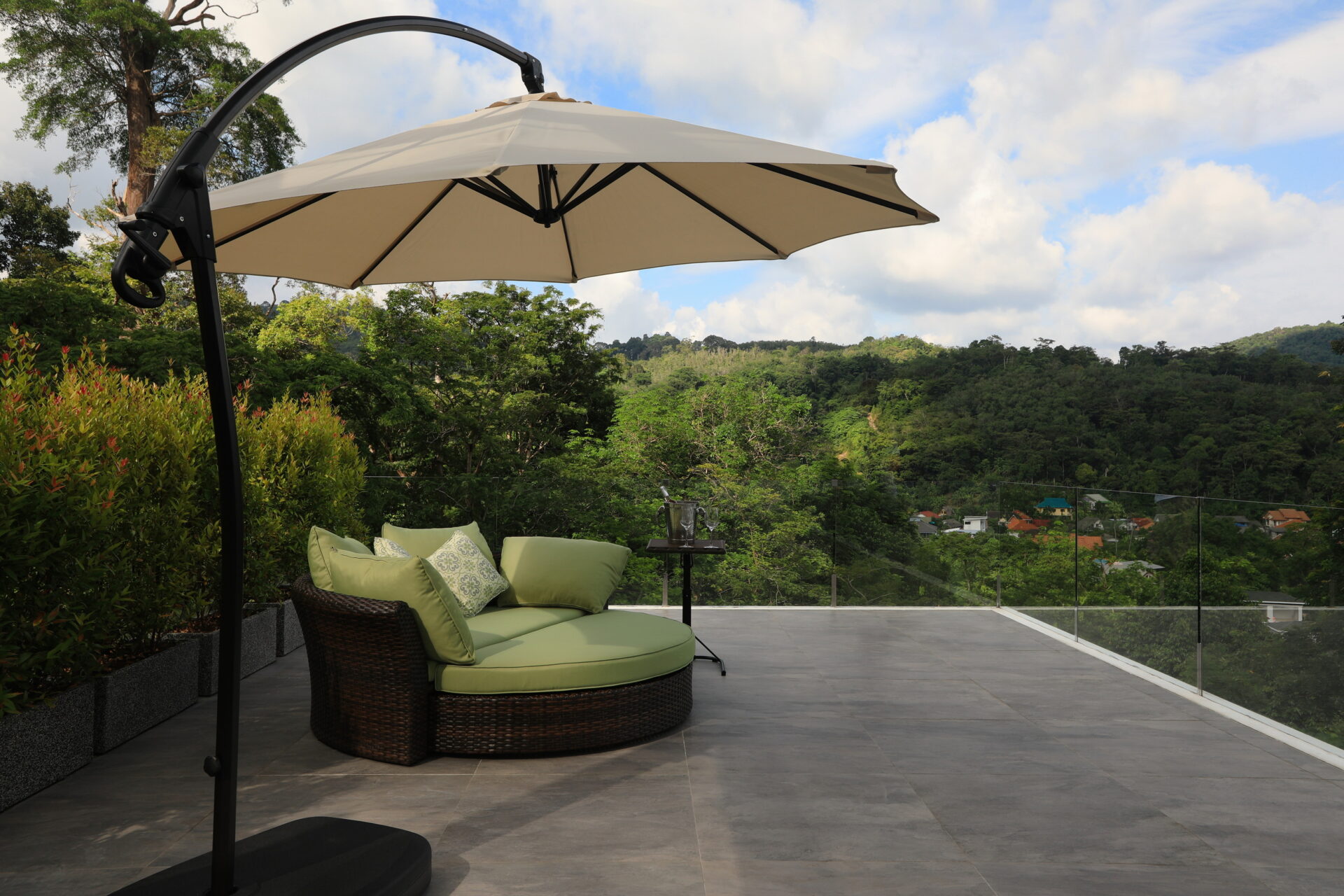 Outdoor terrace featuring a circular wicker sofa with green cushions under a beige cantilever umbrella, overlooking hills and a blue sky.