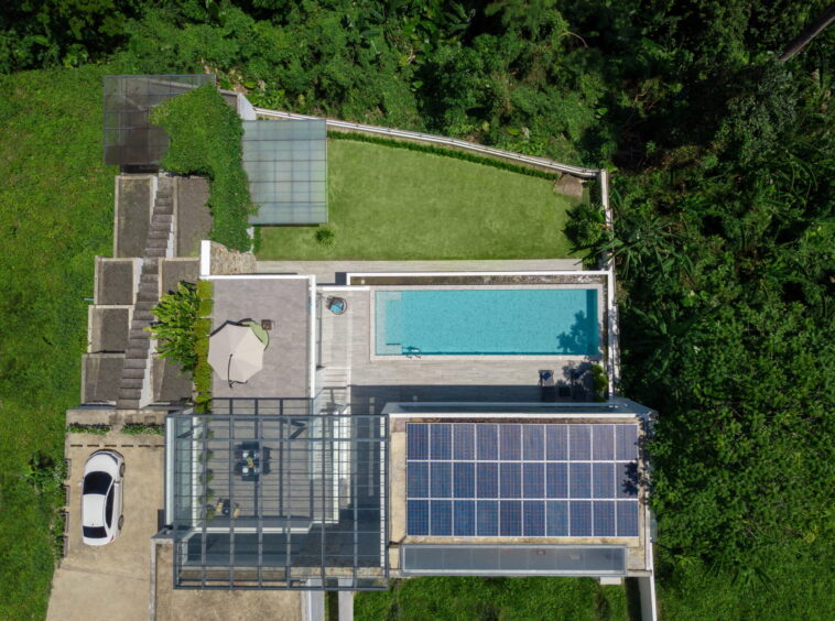 Aerial view of a modern house with a rectangular blue pool, glass conservatory, and surrounding tropical garden.