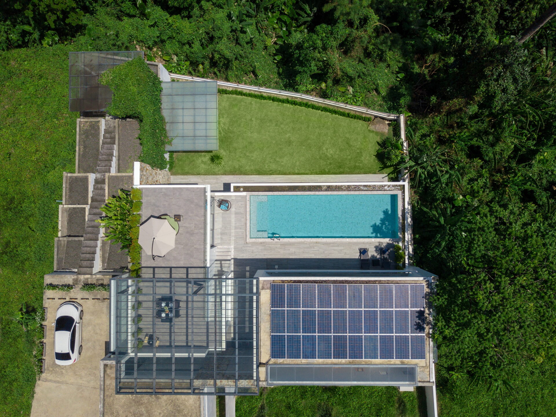 Aerial view of a modern house with a rectangular blue pool, glass conservatory, and surrounding tropical garden.