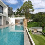 Modern white villa with large glass walls and a rectangular swimming pool, overlooking a green hillside.