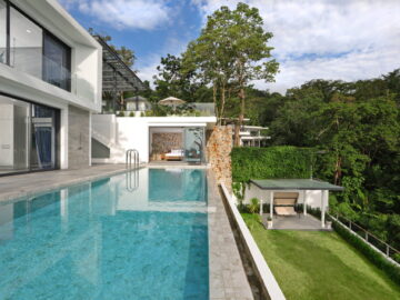 Modern white villa with large glass walls and a rectangular swimming pool, overlooking a green hillside.