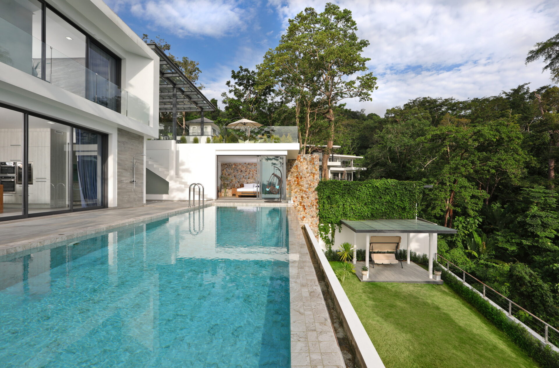 Modern white villa with large glass walls and a rectangular swimming pool, overlooking a green hillside.