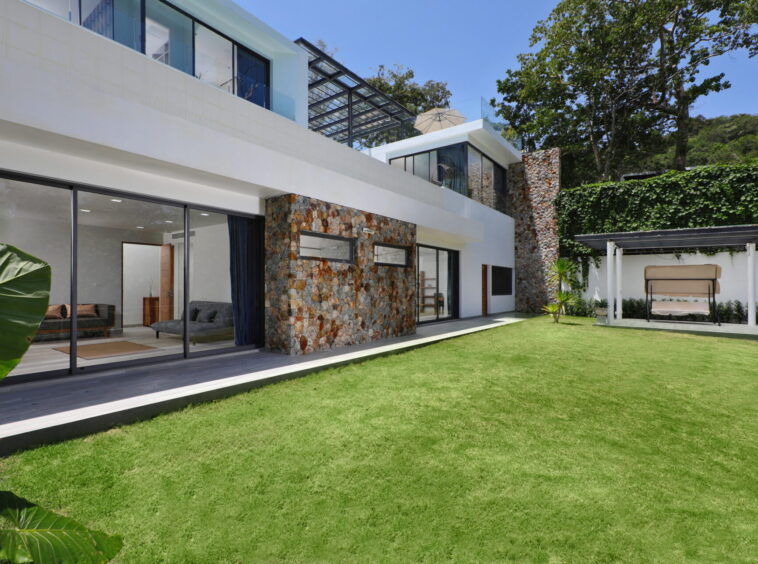 Modern house with glass walls and stone accents opening to a manicured lawn.