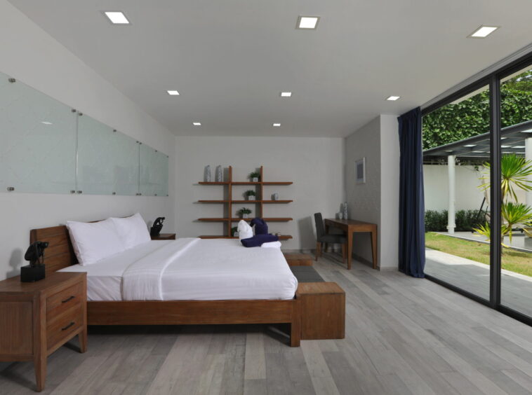 Twin beds with white linens and wooden frames sit side by side in a bright moderne hotel room, with a glass divider on the left and a large glass wall opening to a garden outside.