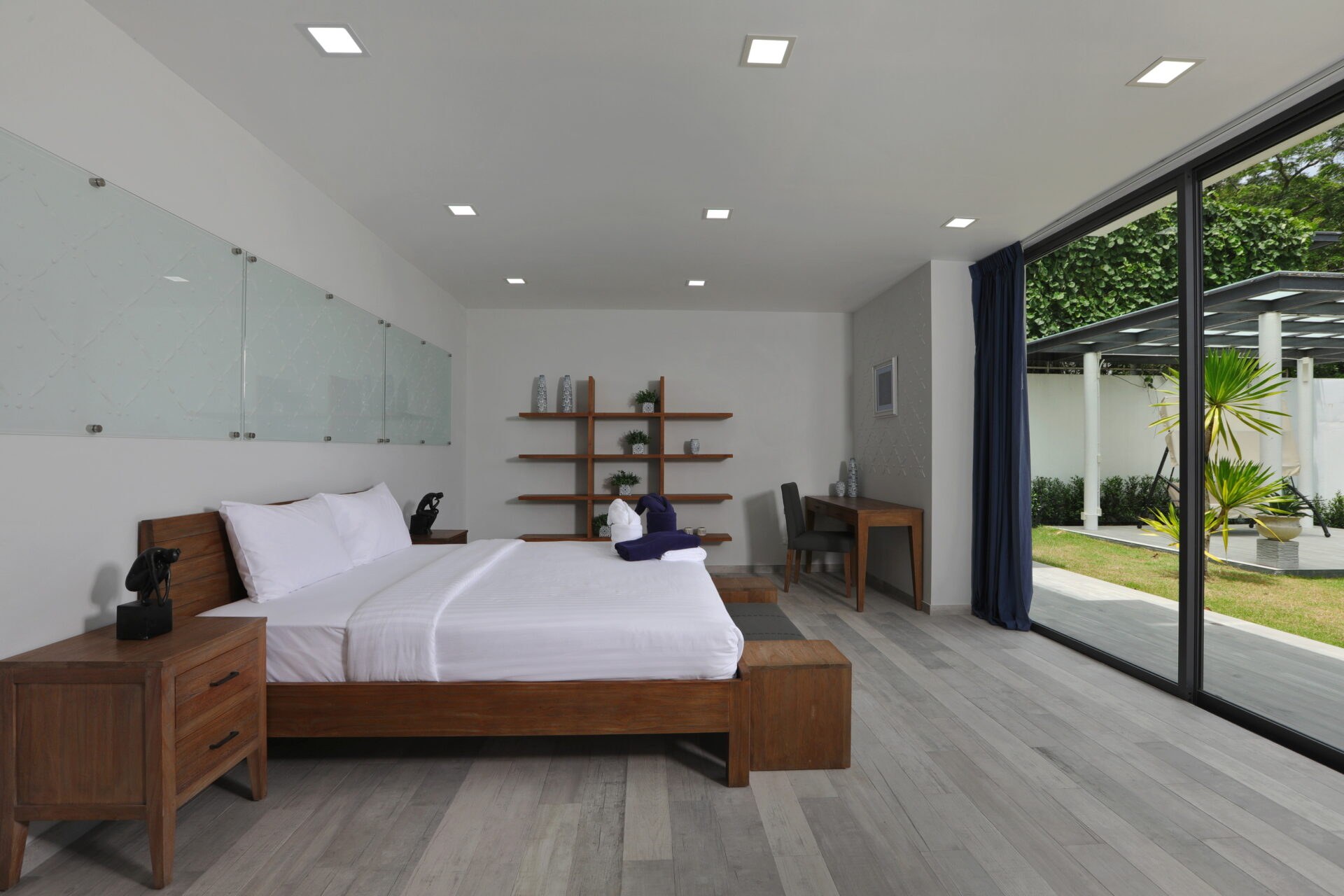 Twin beds with white linens and wooden frames sit side by side in a bright moderne hotel room, with a glass divider on the left and a large glass wall opening to a garden outside.