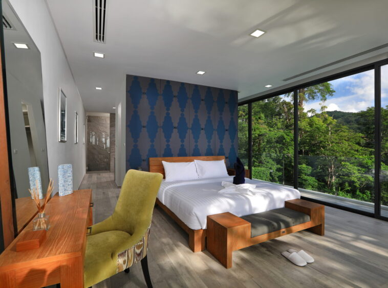Hotel bedroom with floor-to-ceiling windows showing a forest view, a white bed and blue patterned accent wall with wooden furniture.