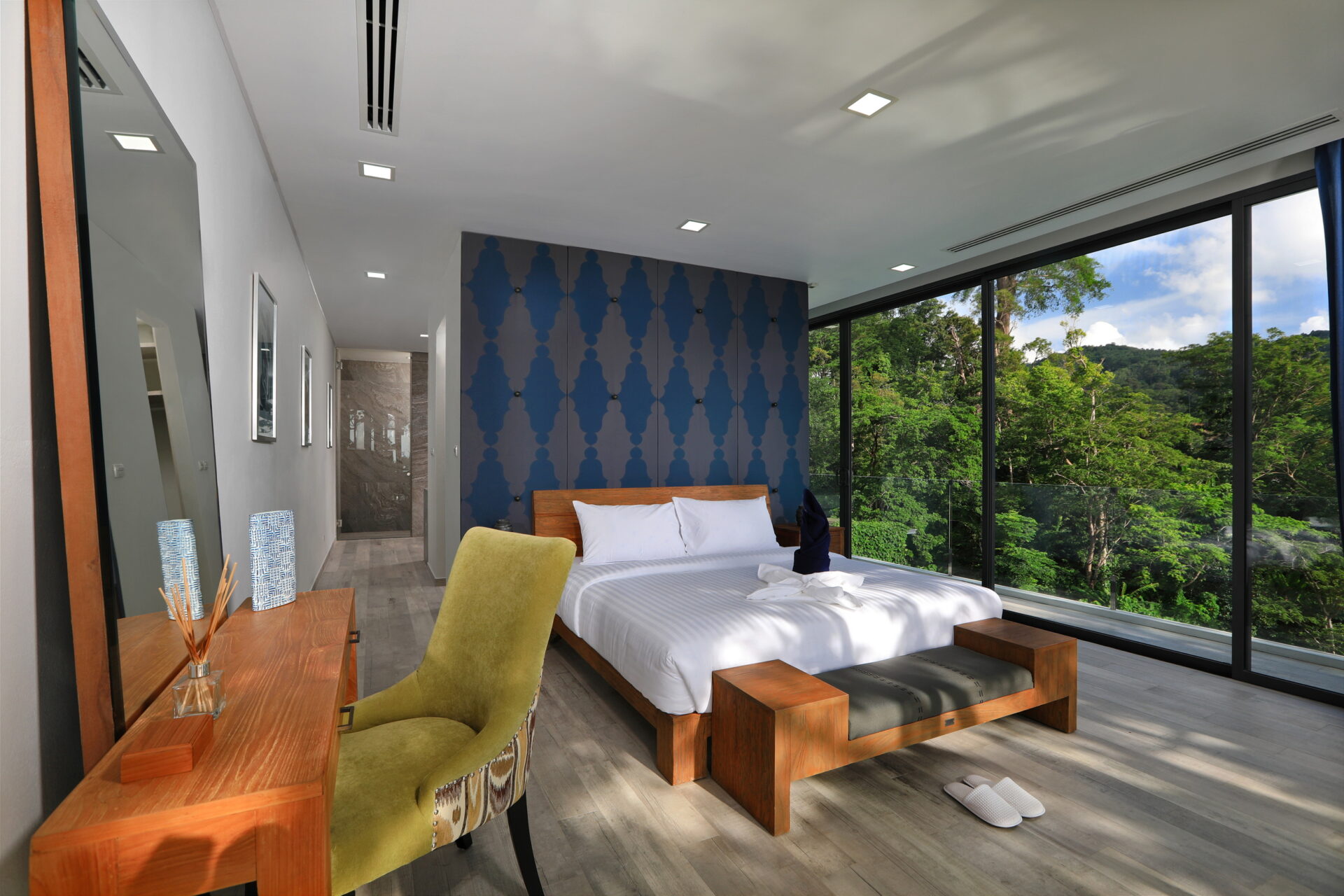 Hotel bedroom with floor-to-ceiling windows showing a forest view, a white bed and blue patterned accent wall with wooden furniture.
