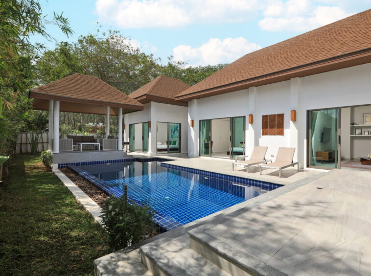 Modern villa with white walls, brown tiled roofs, and a rectangular blue-tiled pool with lounge chairs nearby.