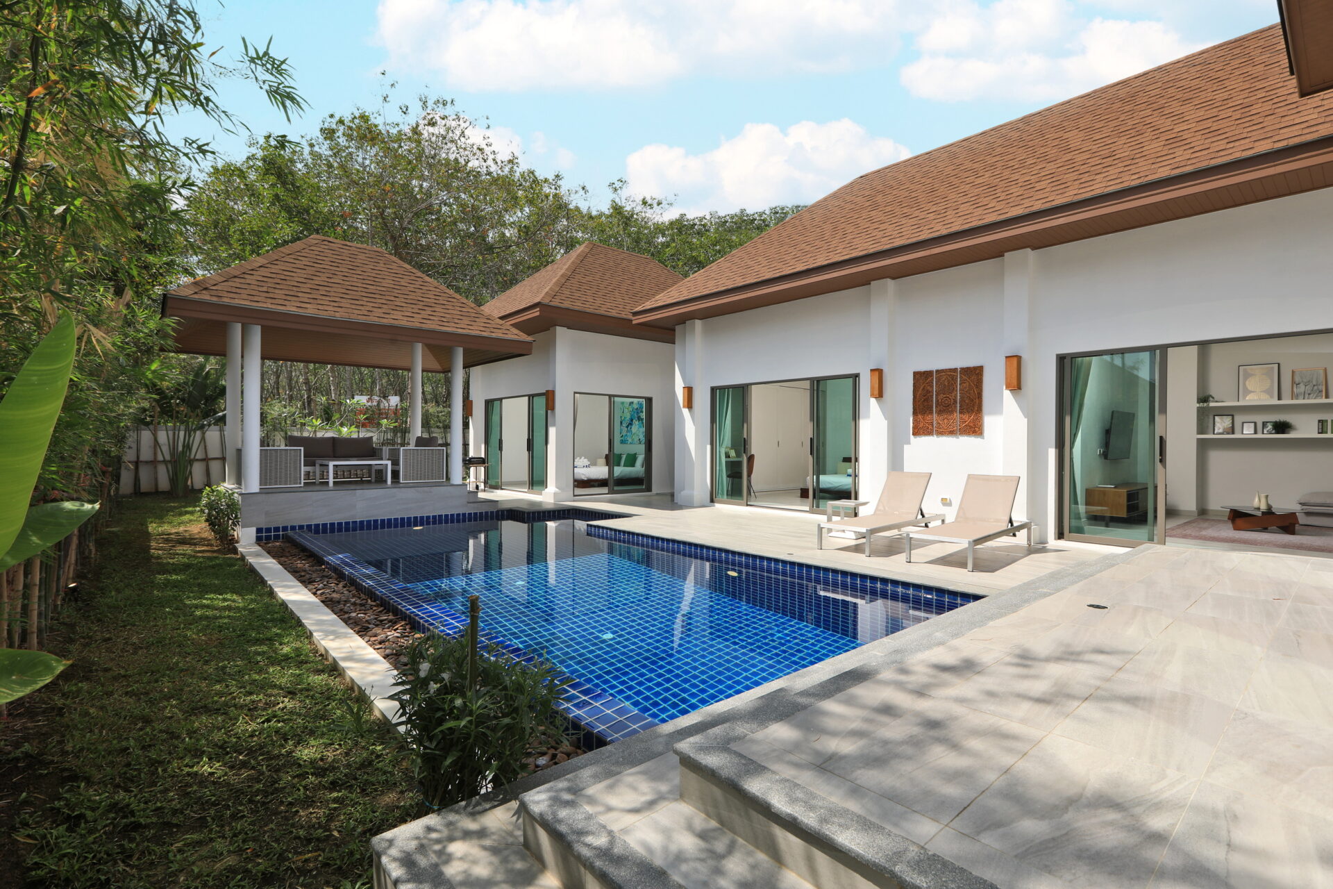Modern villa with white walls, brown tiled roofs, and a rectangular blue-tiled pool with lounge chairs nearby.