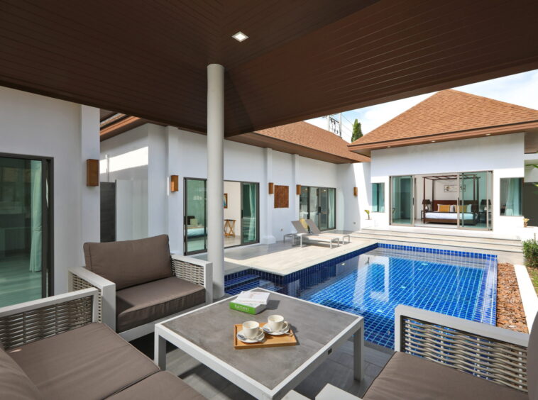 Modern villa courtyard with beige cushioned seating, white furniture, and a blue tiled pool visible through glass doors.