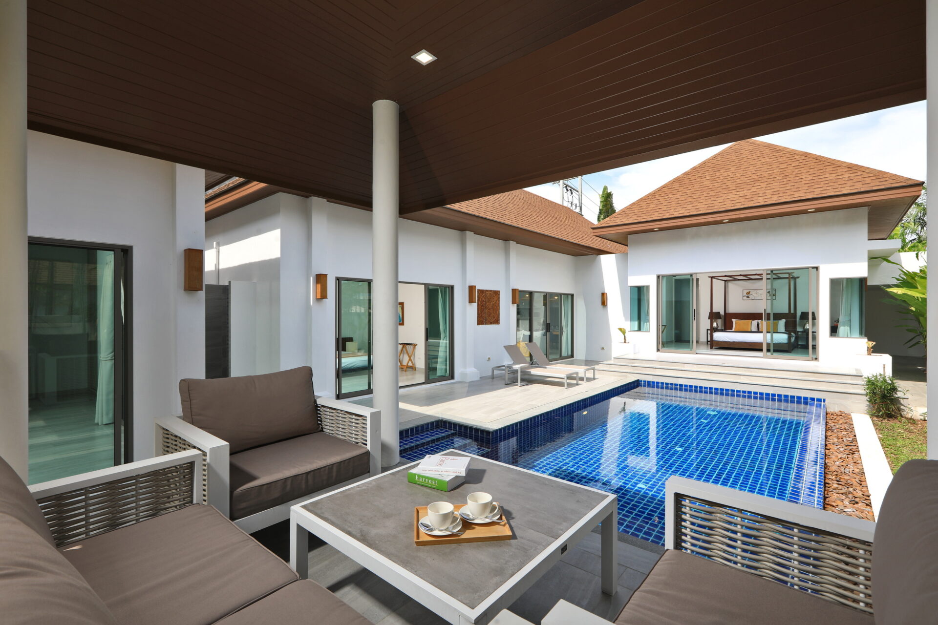 Modern villa courtyard with beige cushioned seating, white furniture, and a blue tiled pool visible through glass doors.