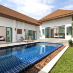 Modern villa with a blue tiled pool, glass sliding doors, and upward-sloped brown roofs; outdoor lounge area.