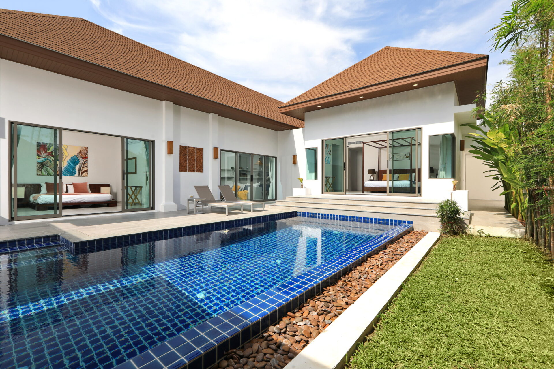 Modern villa with a blue tiled pool, glass sliding doors, and upward-sloped brown roofs; outdoor lounge area.