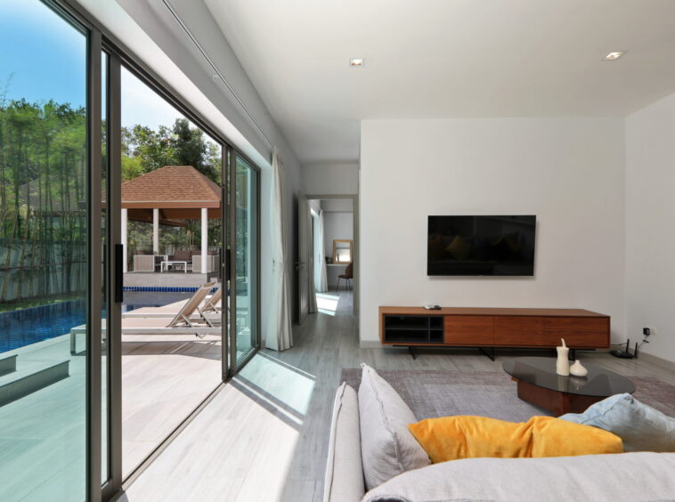Living room with a wall-mounted TV, wooden media console, and gray sofa with orange cushions; large glass doors open to a pool area outside.