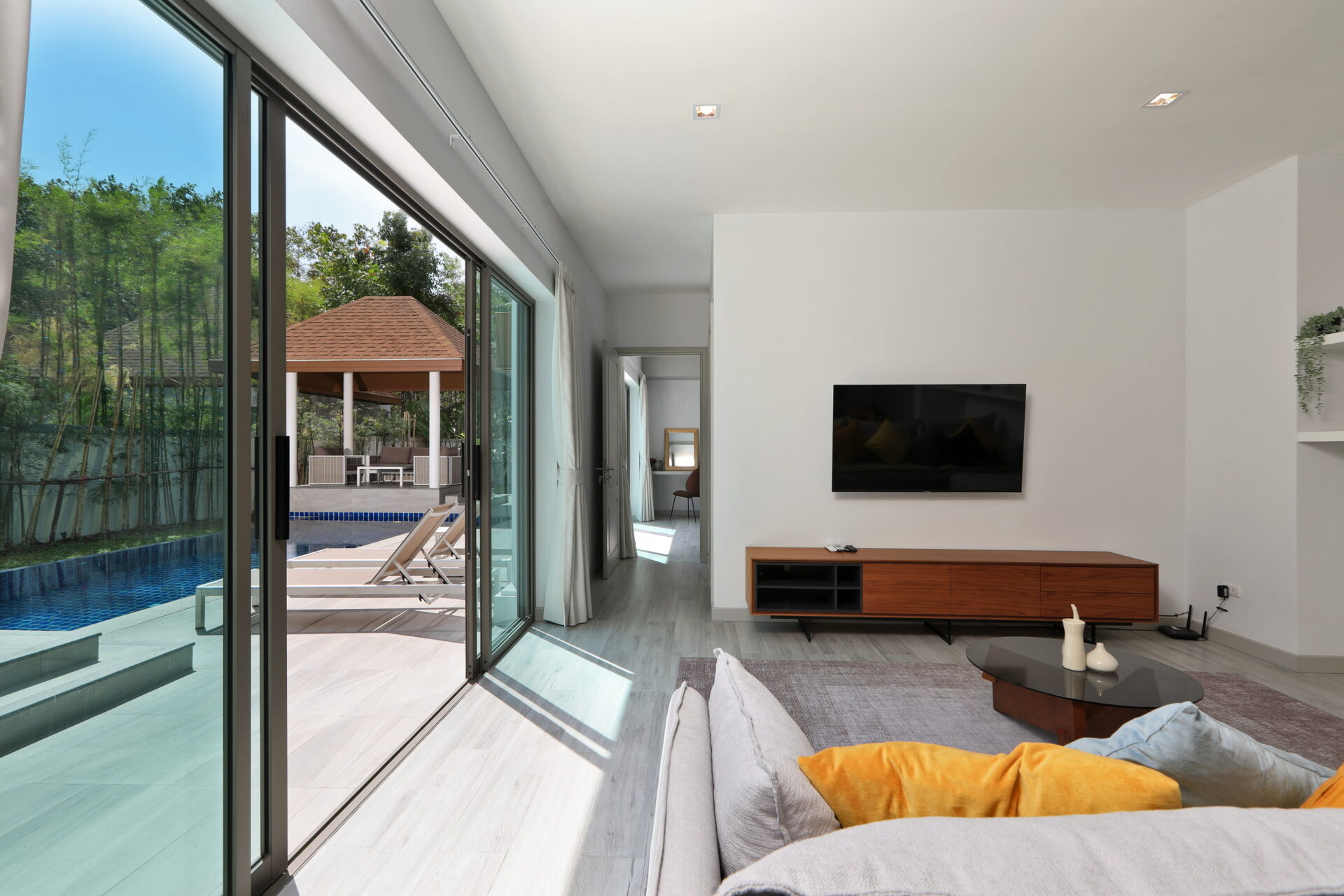 Living room with a wall-mounted TV, wooden media console, and gray sofa with orange cushions; large glass doors open to a pool area outside.