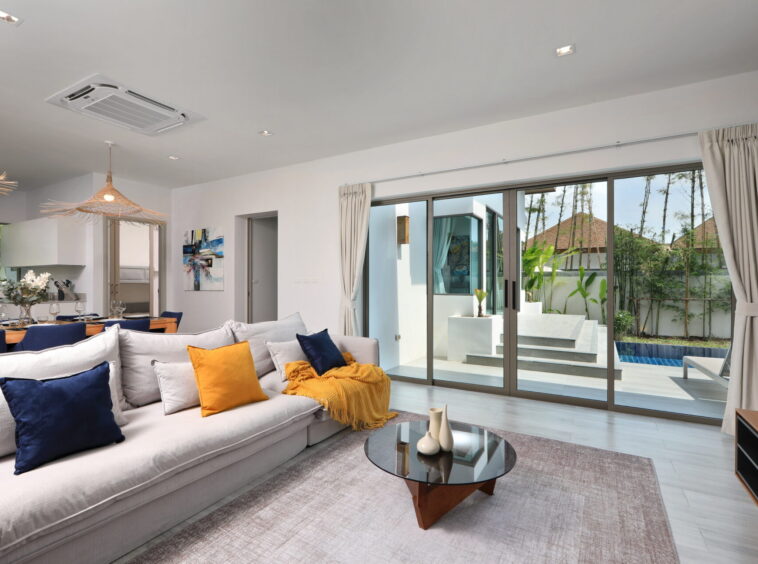 Bright living room with a light-gray sectional sofa, blue and orange cushions, and a glass coffee table, opening to an outdoor patio through large sliding glass doors.