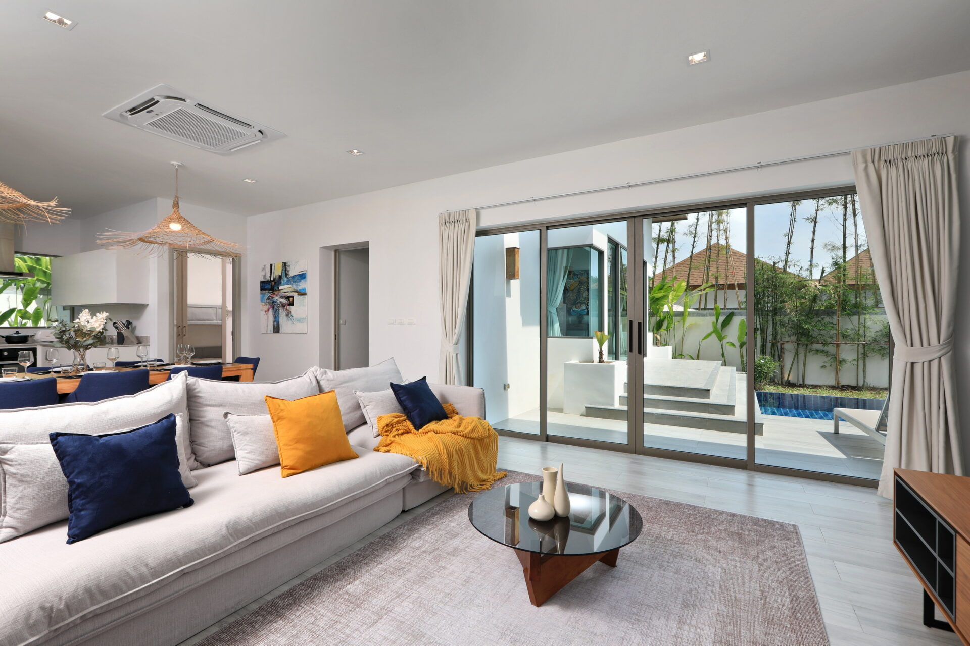 Bright living room with a light-gray sectional sofa, blue and orange cushions, and a glass coffee table, opening to an outdoor patio through large sliding glass doors.
