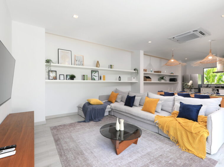 Bright modern living room with a gray sectional sofa, yellow and navy cushions, and a mustard throw; glass coffee table and white shelves with framed art.