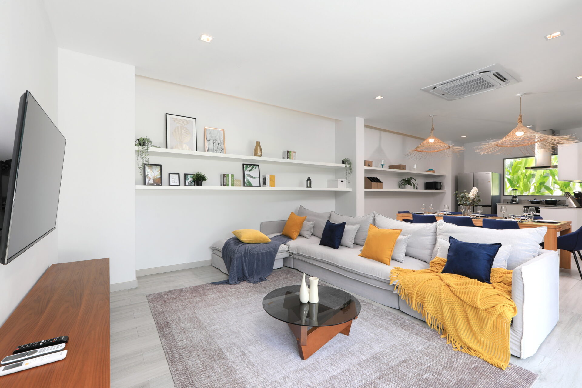 Bright modern living room with a gray sectional sofa, yellow and navy cushions, and a mustard throw; glass coffee table and white shelves with framed art.