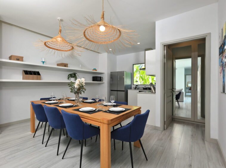Dining area with a wooden table set for eight, blue chairs, and open shelving along a light gray wall beneath woven wicker pendant lights.