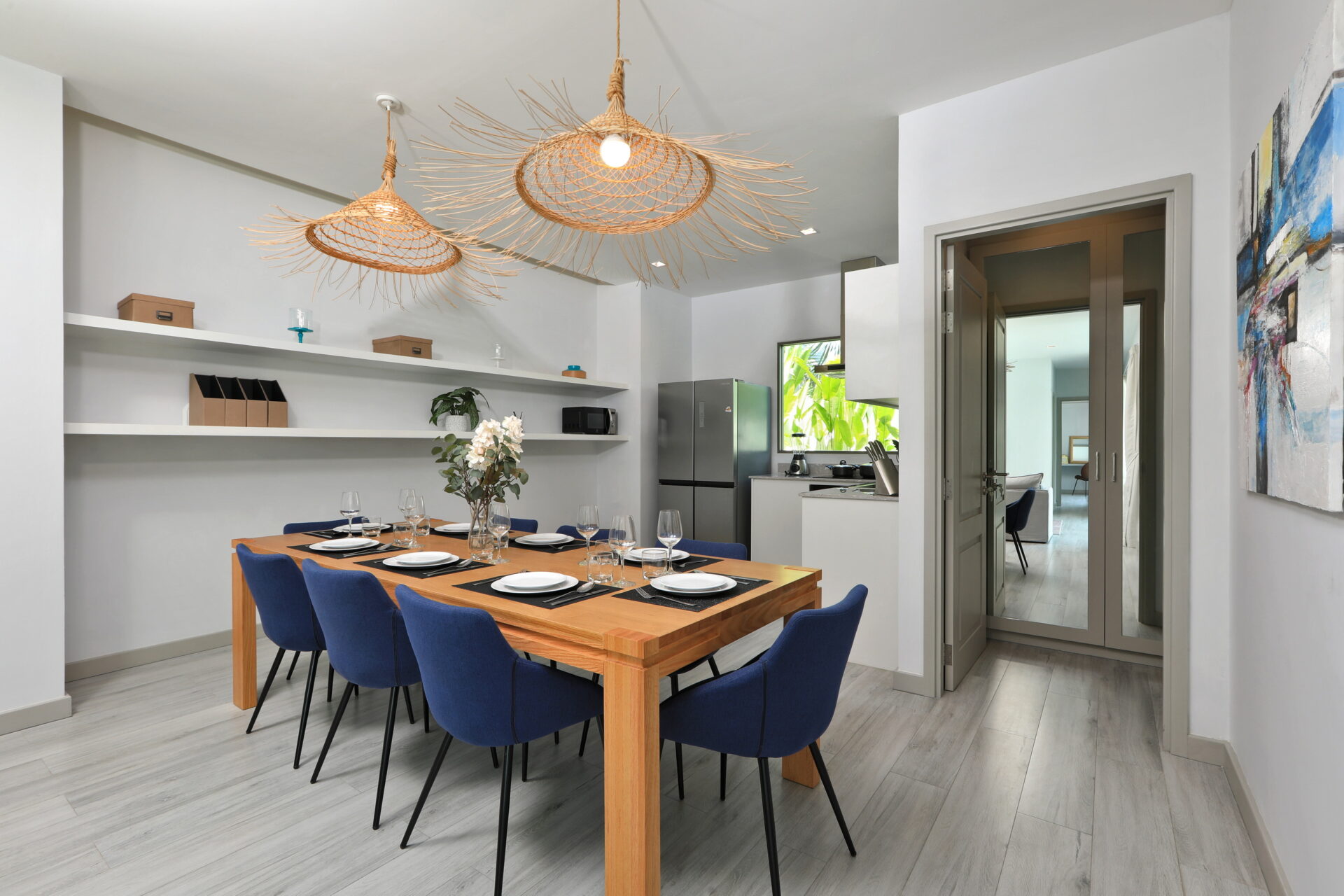 Dining area with a wooden table set for eight, blue chairs, and open shelving along a light gray wall beneath woven wicker pendant lights.