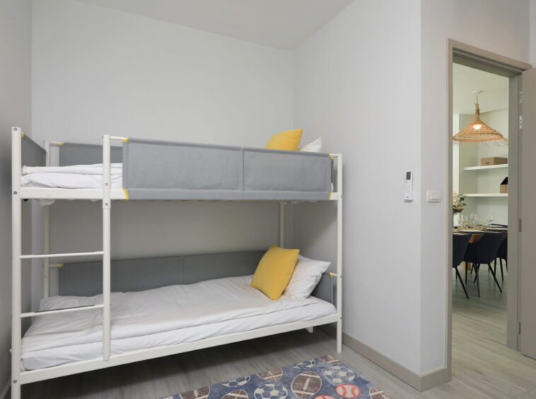 White metal bunk bed with gray fabric panels and yellow pillows in a compact, light-gray bedroom; doorway to a dining area visible.