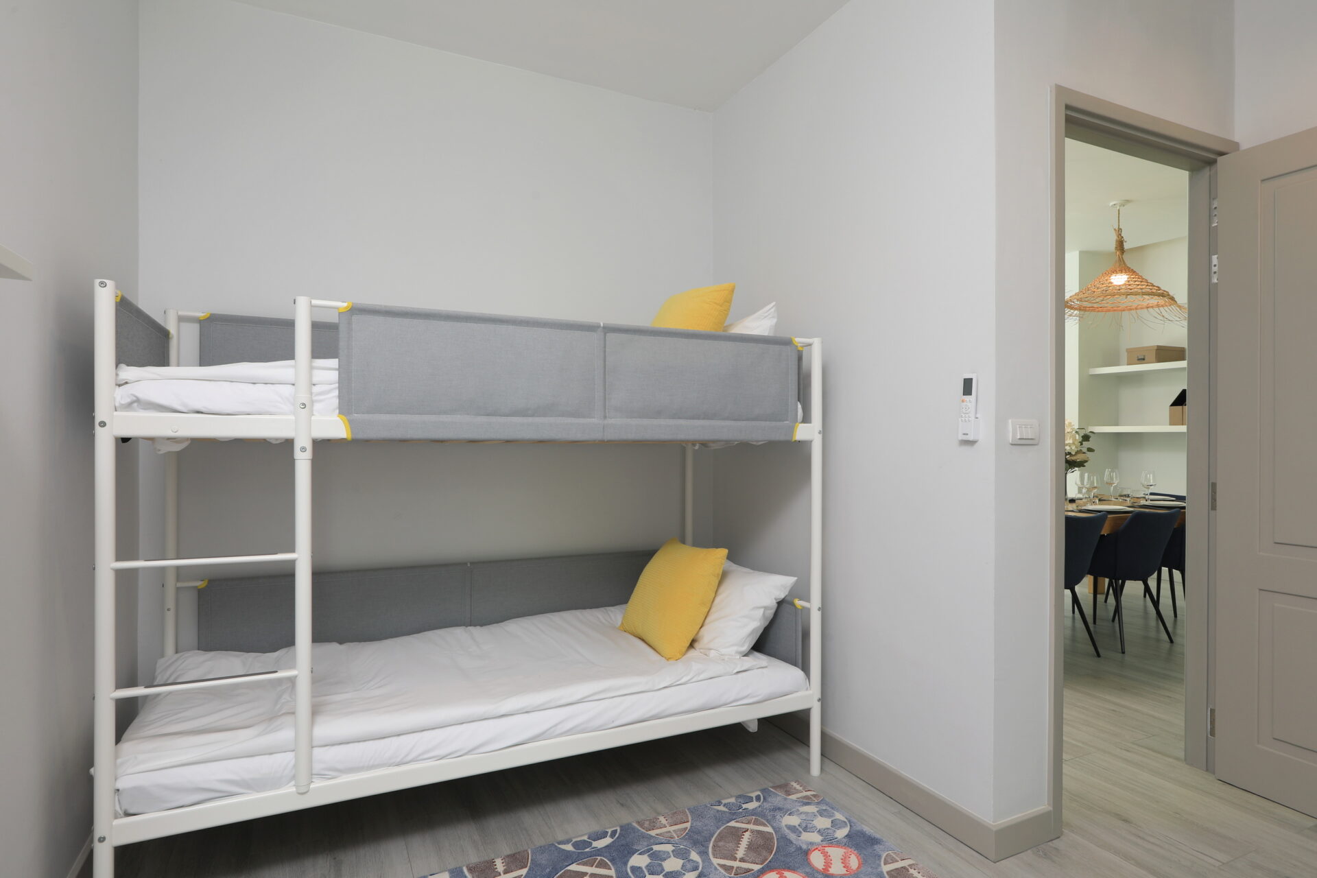White metal bunk bed with gray fabric panels and yellow pillows in a compact, light-gray bedroom; doorway to a dining area visible.