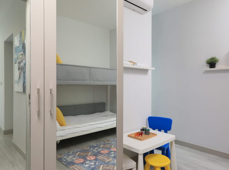 Child’s bedroom with a bunk bed visible through a large mirrored wardrobe, white walls, and a small white table with a blue chair nearby in a bright room.