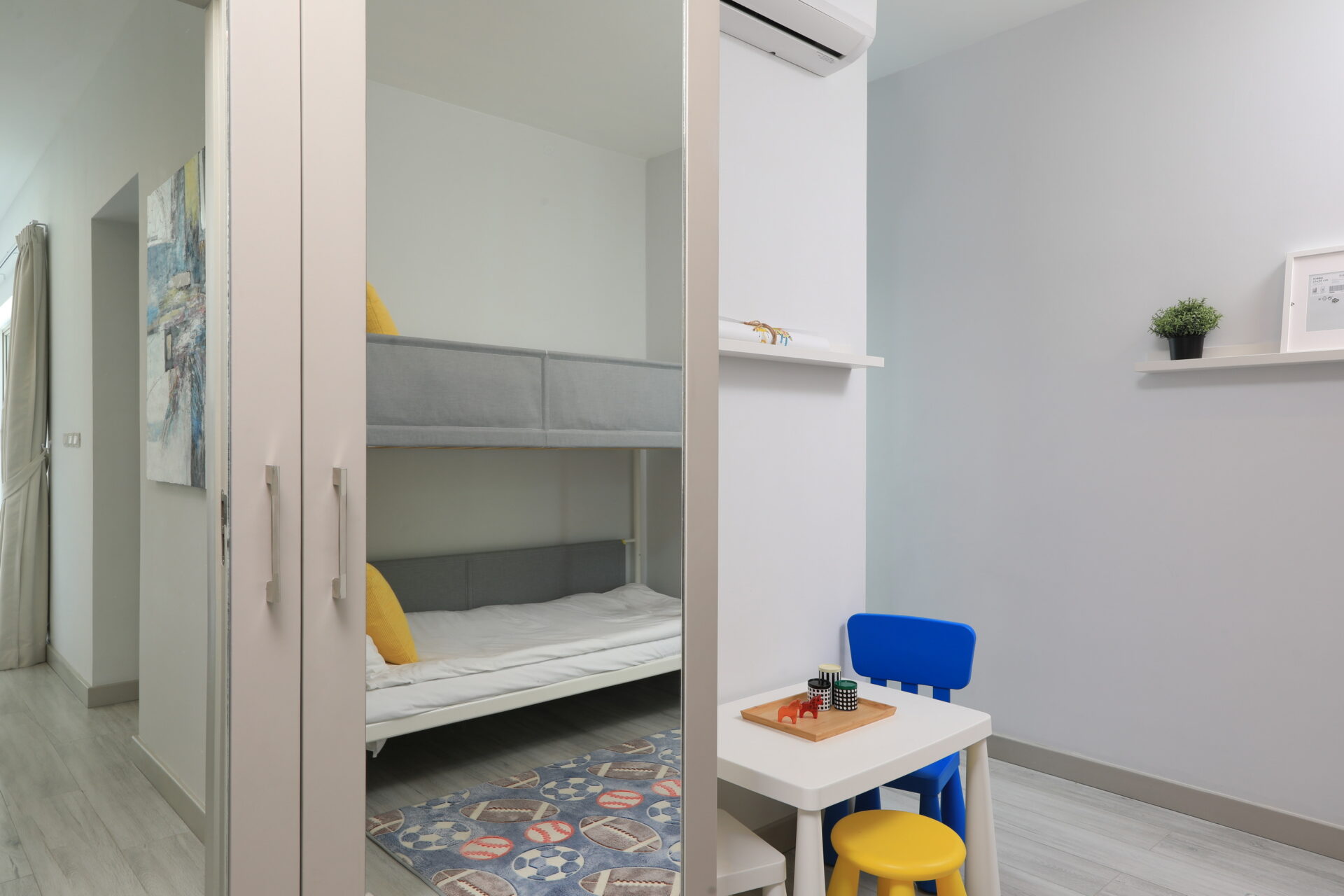 Child’s bedroom with a bunk bed visible through a large mirrored wardrobe, white walls, and a small white table with a blue chair nearby in a bright room.