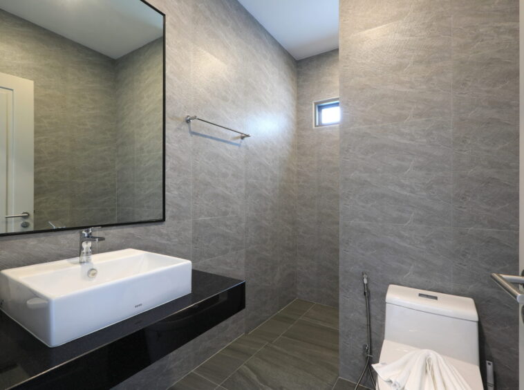 Modern grey-tiled bathroom with a white rectangular sink on a black countertop, large wall mirror, towel bar, and a white toilet.